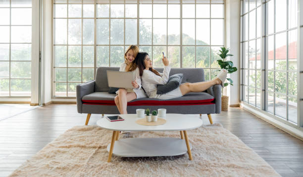 istock Two asian women sit on a couch, one using laptop computer and the other using mobile phone, in a bright room. 2178503378