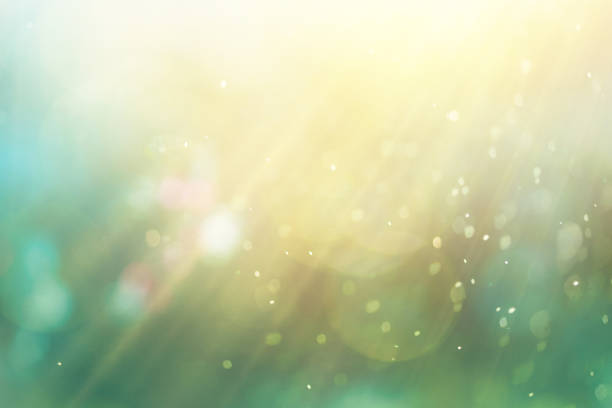 An abstract, ethereal background featuring a defocused design with glowing particles and shiny dust. The scene showcases illuminated elements with a mixture of green and yellow hues, creating a dreamlike quality. Sunbeams filter through, producing light effects and glittering particles that enhance the atmosphere. The textured effect adds depth, while flowing smoke and steam contribute to a sense of mystery. This image is devoid of people and includes ample copy space for text or graphics, making it ideal for celebration-themed designs or decorative purposes.