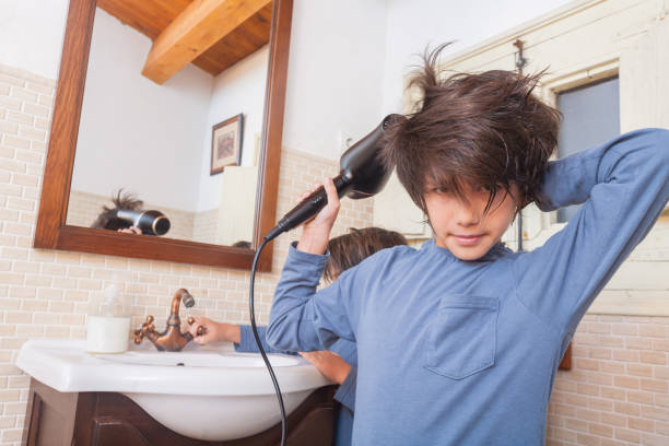 Boy drying his hair with the hairdryer next to the washbasins and mirror in the bathroom.