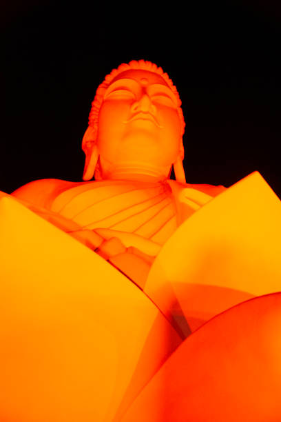 A close-up photograph of a golden statue of a Buddha, illuminated in a warm orange hue. The Buddha's head is tilted upwards and its eyes are closed, giving it a peaceful and serene expression. The black background makes the statue stand out even more.