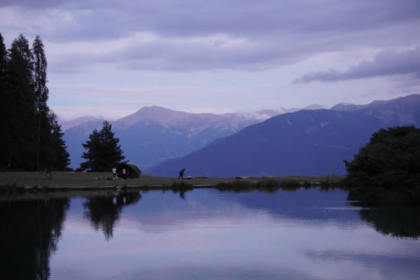 Peaceful camping scene at dusk by an alpine lake, with mountains in the background and serene reflections on the water.