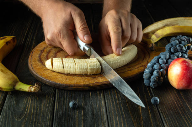 istock Slicing banana with knife in chef hand on kitchen wooden board for preparing fruit dessert. Fruit diet menu 2178519829