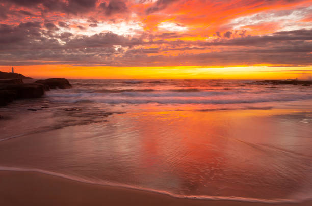 Ocean sunrise with a fiery sky of rich reds and yellows that are reflected in the ocean.  A lighthouse in the far distance