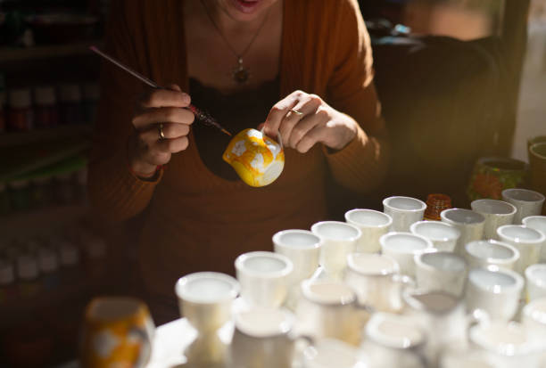 Young female potter decorating handmade ceramic cup using brush and color in pottery workshop.