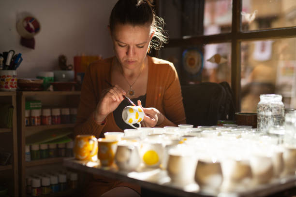 Young female potter painting ceramic mug in art studio.She is making a bunch of beautiful cups to sell in her small pottery shop.