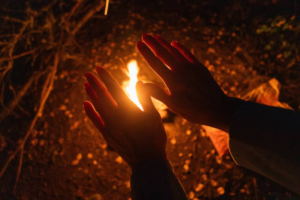Hands are positioned close to a flickering campfire, providing warmth under the night sky. Surrounding darkness highlights the vibrant glow of the fire and offers a serene camping experience.