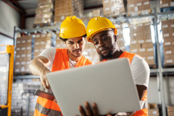 Portrait of two multicultural blue collar workers in working uniforms standing at warehouse with laptop and tracking delivery.