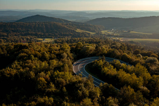 Aerial view of a scenic, winding road in the mountains covered with a forest at sunset.
