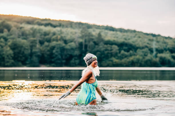 Cold water swimming for elderly women. Senior sporty women standing in lake during cold winter morning, wearing hat and gloves. Cold water swimming for elderly women. Senior sporty women standing in lake during cold morning, wearing hat and gloves. woman cold water swimming stock pictures, royalty-free photos & images