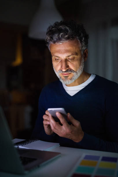 istock Handsome man working from home office at evening, holding smartphone. Screen glow illuminates man's face. Telecommuting and home office. 2178575751