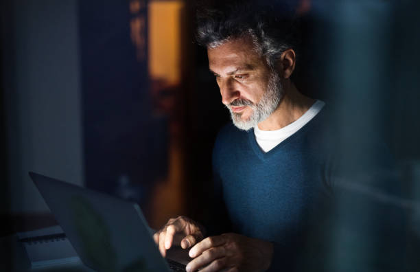 istock Handsome man working from home office at evening, using laptop. Screen glow illuminates man's face. Telecommuting and home office. 2178576416