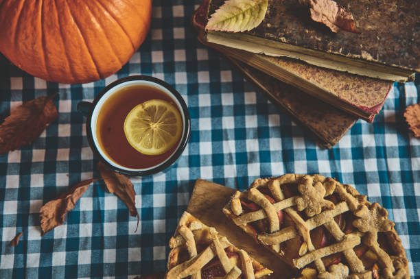 istock Autumn table setting with pumpkin, pie, and tea on checkered tablecloth surrounded by fallen leaves and vintage books, creating a cozy fall atmosphere perfect for seasonal gatherings and relaxation. 2178580927