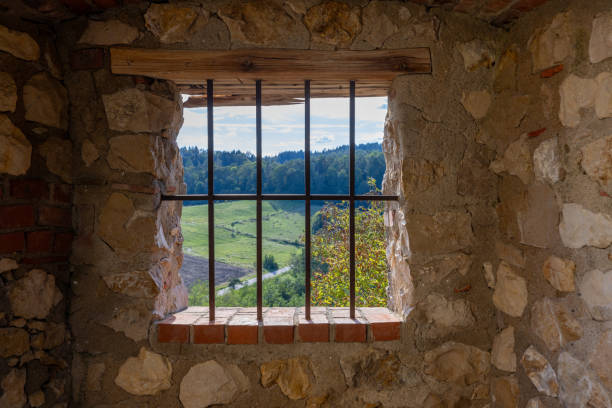 A barred window in a stone wall frames a peaceful view of green hills, fields, and distant forests during daytime