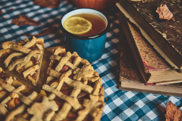 A delicious apple pie with lattice crust served alongside a cup of lemon tea resting on a checkered tablecloth, with vintage books in the background, radiating warmth and coziness.