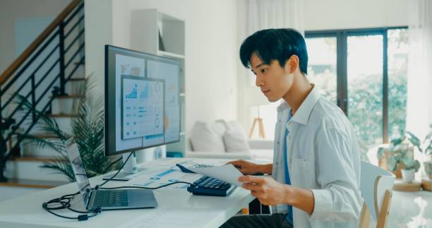 istock Young Asian male working from home, analyzing financial data on computer screens, reflecting modern home office setup and remote work. Remote work and finance. 2178582913