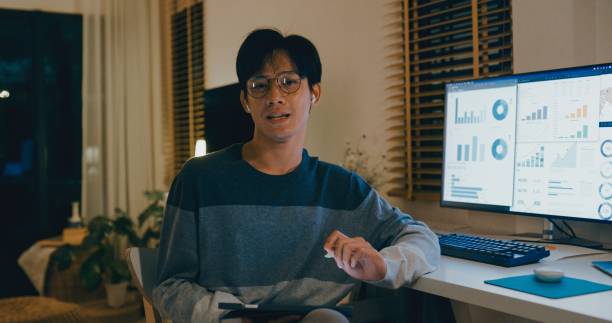 Young Asian man financial advisor working late at home, using tablet and laptop, discussing strategy with clients via video call, showcasing remote work efficiency. Remotely and finance.