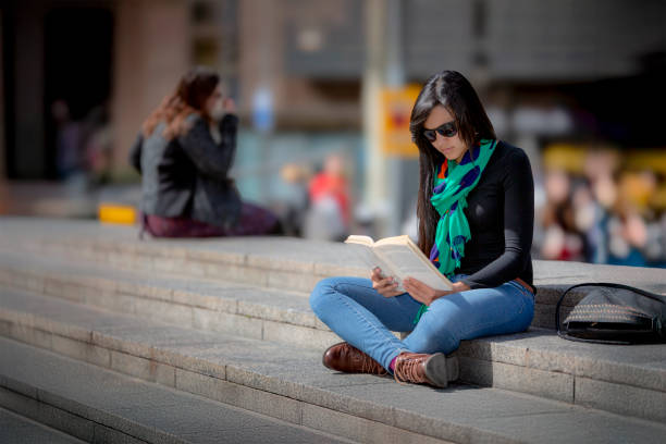 Young university woman reading a book sitting on a stone bench in her break surrounded by a beautiful green grass