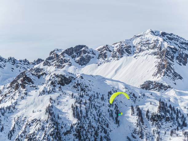 A paraglider with a vibrant orange wing soars over snow-covered mountains at Serre Chevalier, Briançon, France. The bright sky and rugged peaks create a stunning alpine winter landscape.