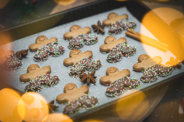 Three rows of gingerbread men with chocolate and sprinkles on white baking paper in a baking dish with cinnamon, candy cane, fir branches, golden bokeh and selective focus on a dark cement backdrop, close-up side view. Holiday food concept.