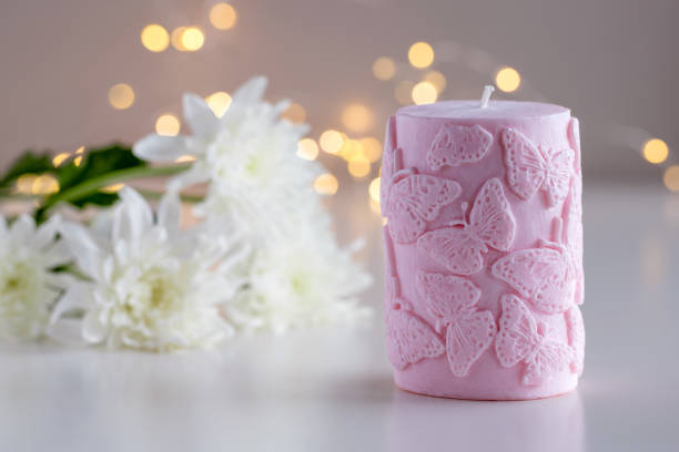 Pink decorative candle with butterflies pattern. Flowers and lights in background with copy space. Home decor. Cozy interior. Soy wax candle making.