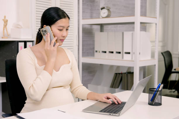 Pregnant Asian woman holding phone to ear while working on laptop in home office environment promoting work-life balance and multitasking during pregnancy lifestyle concept. mujer asiática embarazada que sostiene el teléfono a la oreja mientras trabaja en una computadora portátil en un entorno de oficina en casa que promueve el equilibrio entre el trabajo y la vida personal y la multitarea durante el concepto de estilo de v