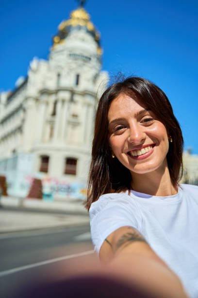 Young woman taking a selfie with historic building in Madrid Portrait of a young woman taking a selfie, smiling broadly, with a historical building and clear blue sky in the background. selfie madrid photos stock pictures, royalty-free photos & images