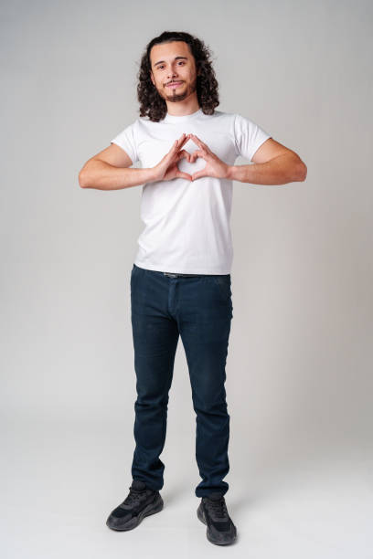 istock Young man with curly hair forms a heart shape with his hands while standing against a light background 2178663593