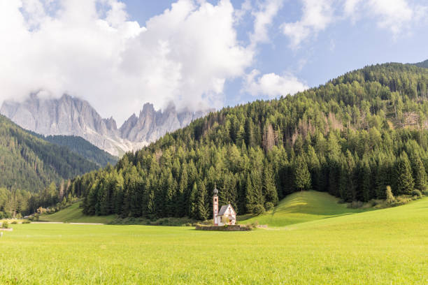 A charming small church sits peacefully in a lush green meadow, framed by dense forests and towering mist-covered alpine peaks.