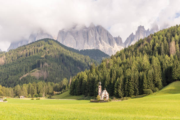 A charming small church sits peacefully in a lush green meadow, framed by dense forests and towering mist-covered alpine peaks.