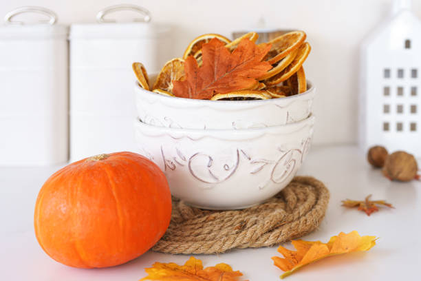White bowl filled with dried orange slices, orange pumpkin and colorful autumn leaves on background in cozy kithen. Home fall decor, beautiful tableware