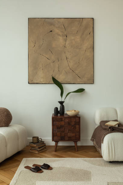 istock A modern living room with a white sofa, wooden side table, and neutral decor. An abstract painting hangs above the table, which holds a vase with green leaves and a bowl. Books, slippers, and a coffee cup add a cozy touch. 2178686525