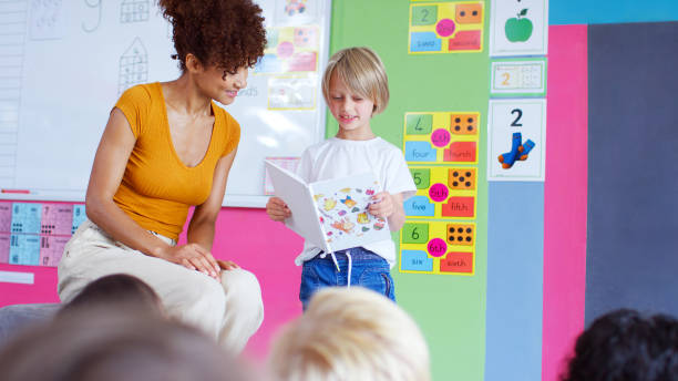 istock Male Pupil In Elementary School Classroom Reading Book To Class With Teacher 2178694157