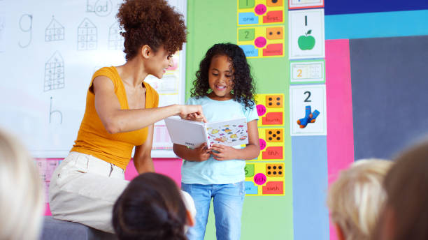 istock Female Pupil In Elementary School Classroom Reading Book To Class With Teacher 2178694173