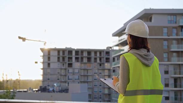 istock Female construction engineer with a hard hat and safety vest is writing on a clipboard while inspecting a construction site in the evening at sunset 2178699866