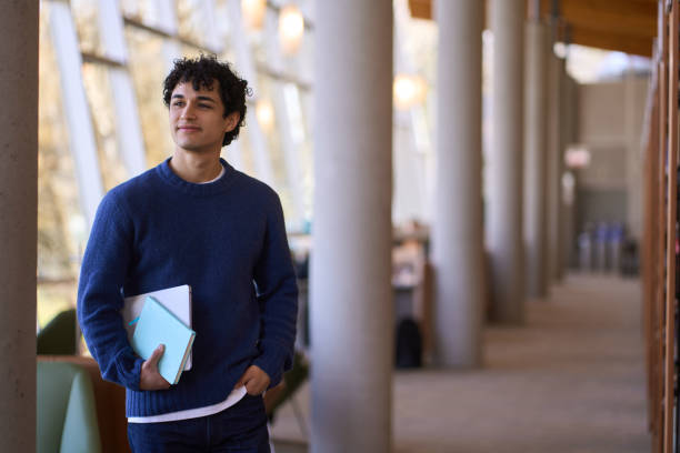 istock Young student walking in university hall holding books and laptop looking away 2178702843