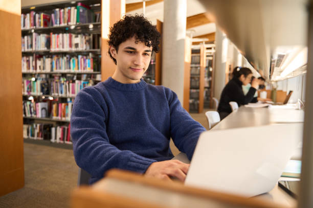 istock Young man working on laptop sitting at desk in busy public library 2178703008