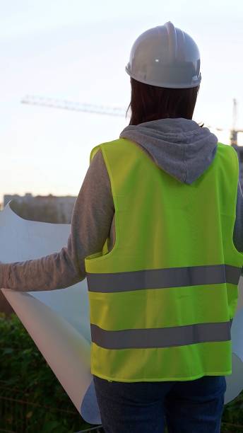 istock Woman an architect or engineer wearing safety hard hat and vest holding blueprint while inspecting a construction site at sunrise, vertical back view. Architecture and engineering 2178706492