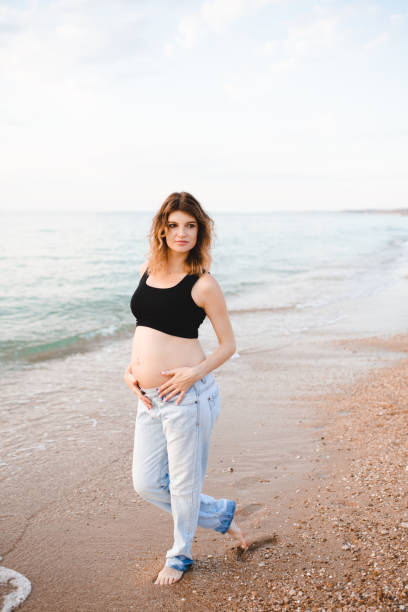 Pregnant woman 29-30 year old resting at beach over open sea wearing denim pants and top. Motherhood. Summer vacation time.