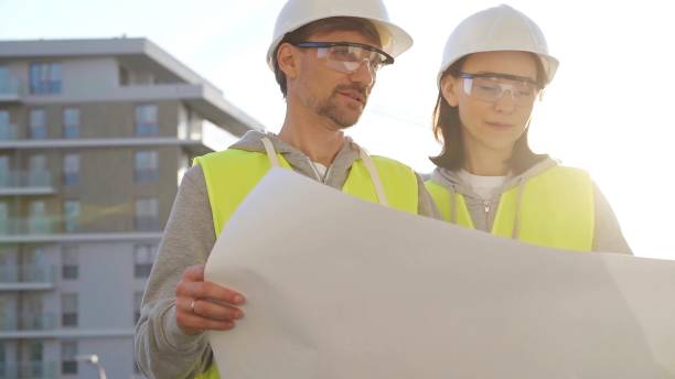 istock Architects or engineers wearing safety helmets and vests holding blueprint and discussing something on construction site at sunrise, front view. Architecture and engineering 2178719169