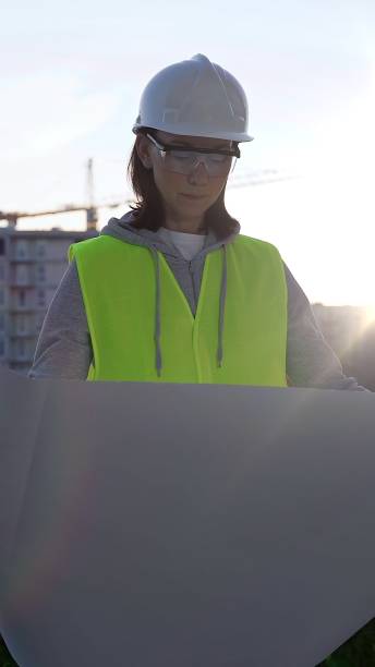 istock Female architect or engineer wearing safety hard hat and vest holding blueprint while inspecting a construction site at sunrise in early morning, front vertical view. Architecture concept 2178719354