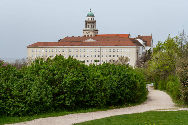 A view of the Benedictine Pannonhalma Archabbey, showcasing its iconic dome and red-tiled roof, framed by a lush garden pathway. The historic monastery stands as a symbol of Hungarian religious and cultural heritage, surrounded by greenery and a peaceful landscape.