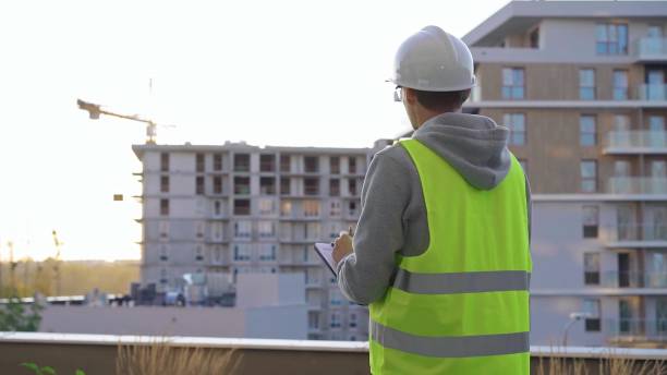 istock Male constructive engineer with a white protective helmet and safety vest is writing on a clipboard while inspecting a construction site at sunset. Architect concept 2178732061