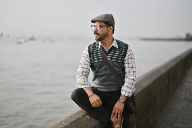 A mid-adult man sits alone at a peaceful seafront in Mumbai, gazing out over the vast expanse of the sea, casually dressed in a cozy sweater, wearing glasses, and carrying a sling bag.