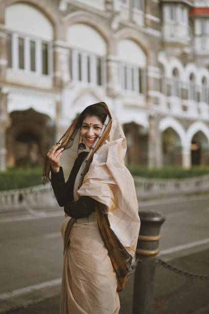 A woman with her sari pallu elegantly draped over her head smiles warmly while gazing directly at the camera. Her expression exudes a gentle warmth and confidence, capturing a timeless sense of grace. The colors and texture of her sari add cultural richness to the scene, highlighting the elegance and beauty of traditional attire.