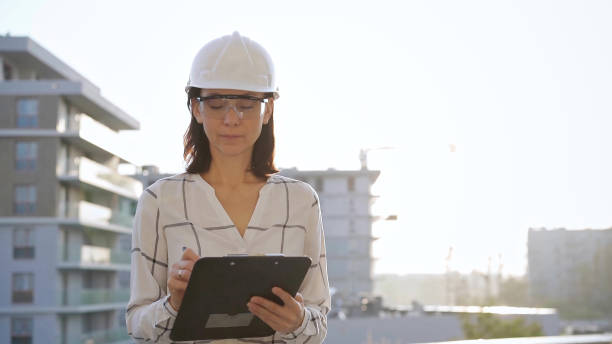 istock Woman construction engineer wearing white checked blouse and hard hat is making notes on a clipboard while inspecting a building site at sunset, front view. Architecture and engineering concepts 2178757273