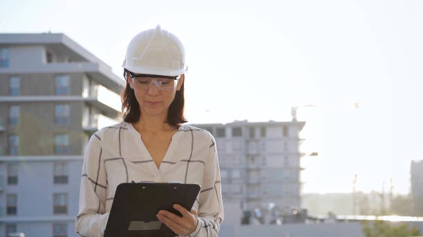 istock Woman construction engineer wearing white checked blouse and hard hat is making notes on a clipboard while inspecting a building site at sunset, front view. Architecture and engineering concepts 2178757701