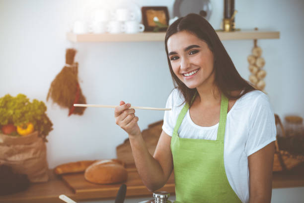 istock An attractive young dark-haired woman tasting ready hot meal with a wooden spoon while standing and smiling in sunny kitchen. Cooking and householding concepts 2178763767