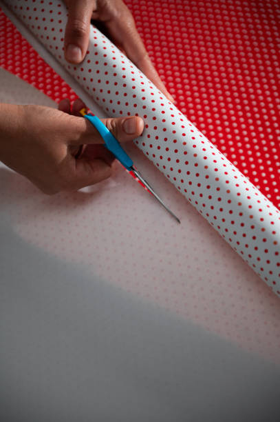 Hands cutting polka dot wrapping paper with blue-handled scissors in a close-up view. The scene captures the holiday gift wrapping process, focusing on precision and creativity during festive preparations.