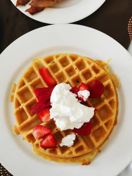 A tempting stack of waffles topped with fresh strawberries and a generous dollop of whipped cream is elegantly displayed on a white plate, set against a sleek black table. Beside it, a second white plate features crispy bacon, creating a mouthwatering breakfast spread.