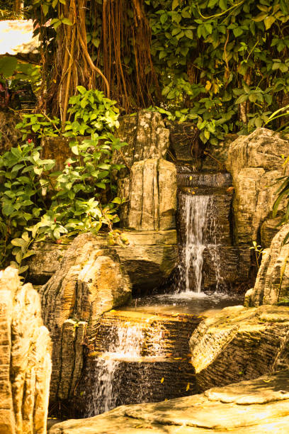 The waterfall flows gracefully over natural rocks in Taman Rimba Melaka, Malaysia, surrounded by vibrant greenery and lush plants, creating a peaceful atmosphere in the forest.